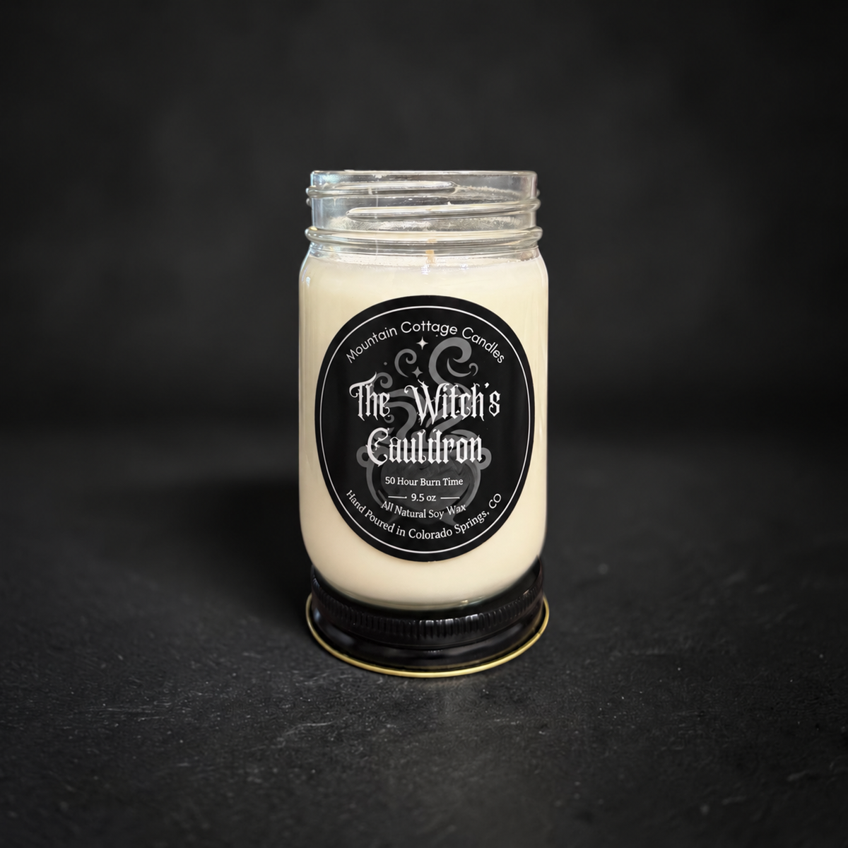 The Witch's Cauldron soy candle in a clear glass jar with a black lid, black label with ornate scrollwork and sparkles