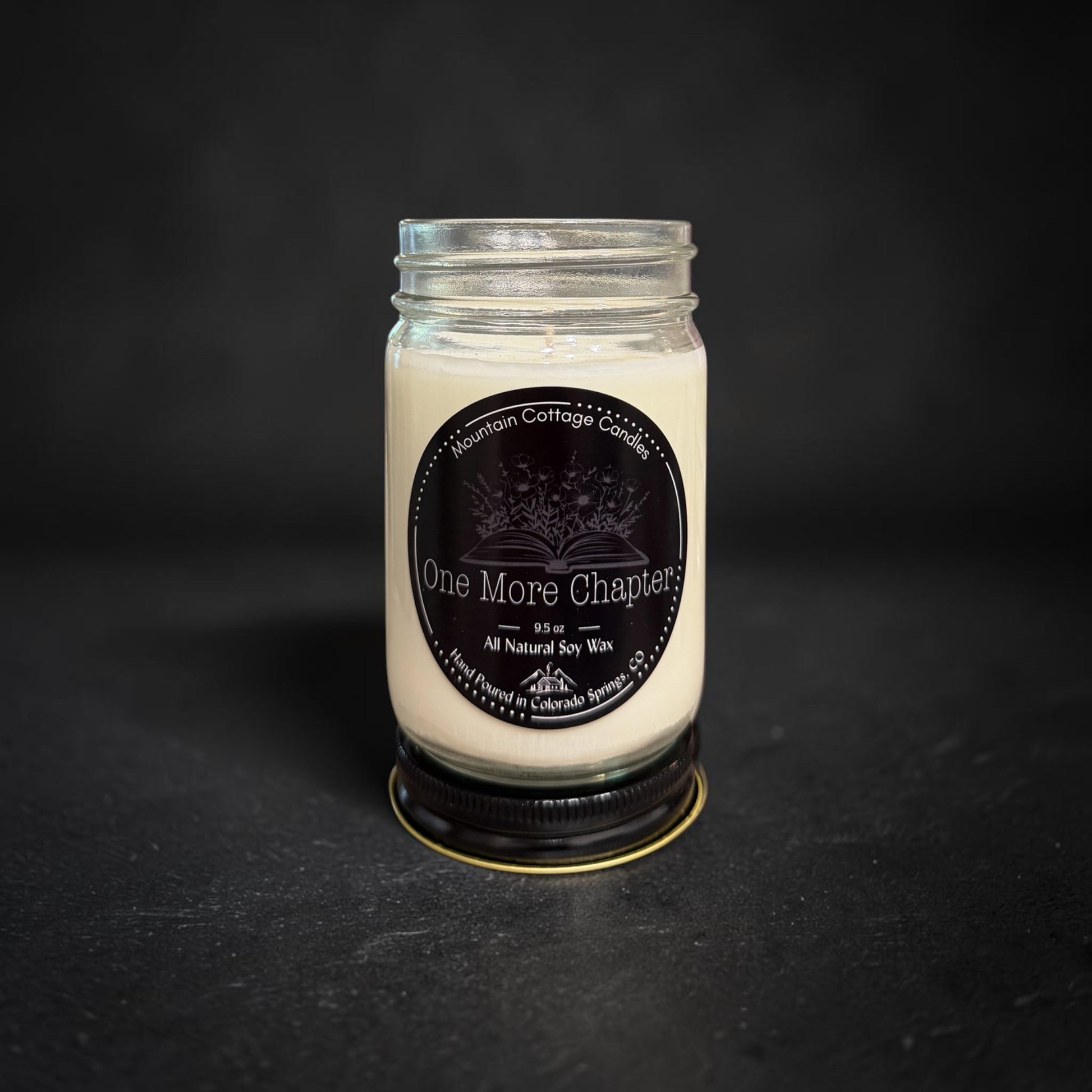 One More Chapter soy candle in a clear glass jar, black label showing an open book with wildflowers rising from its pages