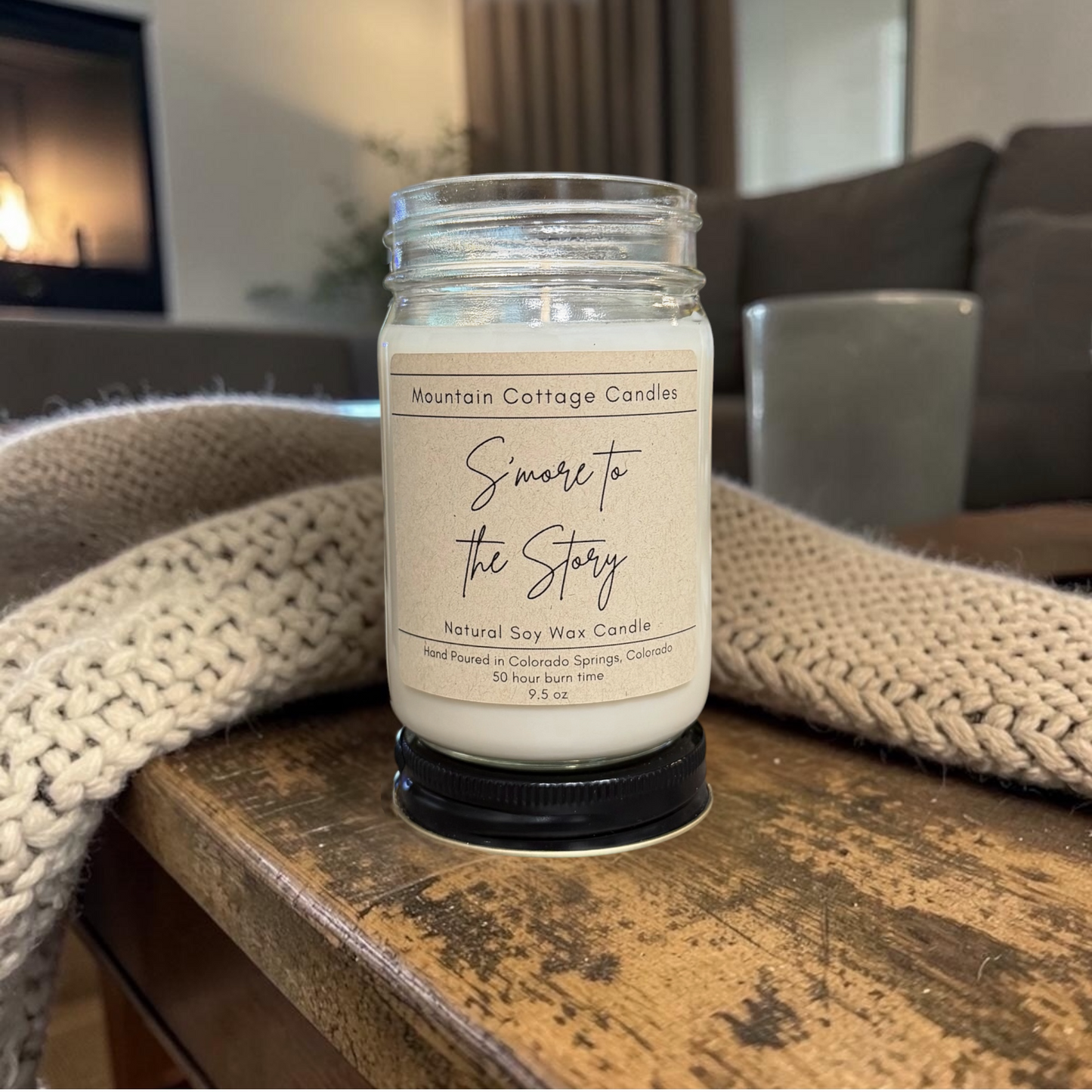 S'more to the Story 9.5 oz soy candle in a kraft-labeled glass jar on a wood side table with a cream knit blanket behind