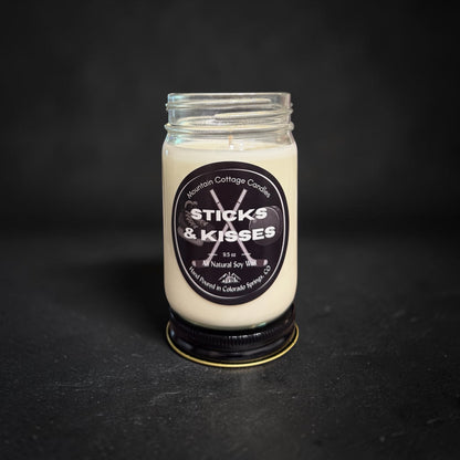 Sticks & Kisses soy candle in a clear glass jar with a black lid, black label showing crossed hockey sticks and a puck