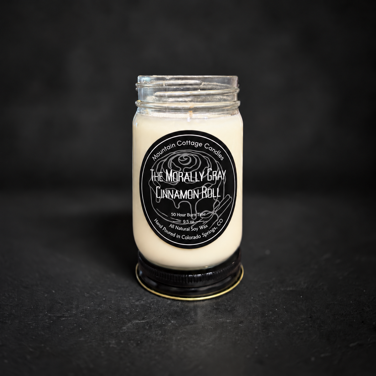 The Morally Gray Cinnamon Roll soy candle in a clear glass jar with a black lid, black label with a line-drawn iced cinnamon roll