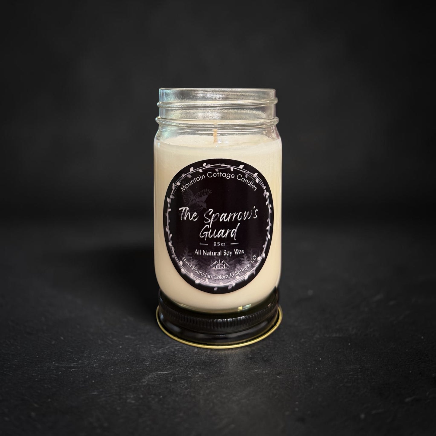 The Sparrow’s Guard soy candle in a clear glass jar, black label showing a sparrow flying above a pine forest