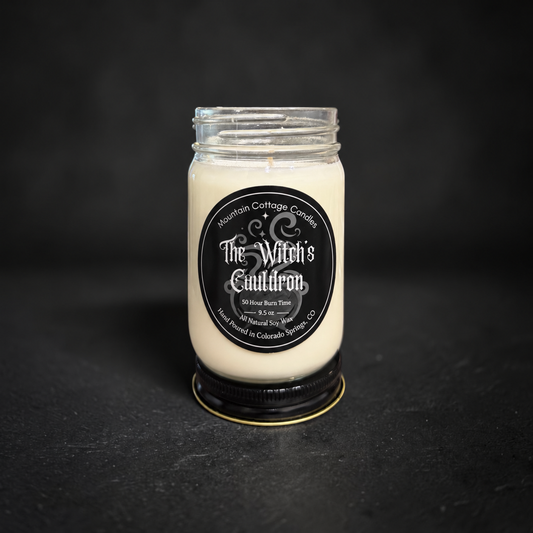 The Witch's Cauldron soy candle in a clear glass jar with a black lid, black label with ornate scrollwork and sparkles